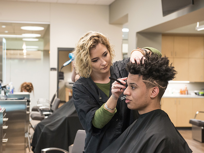 A barbering student shaving the side of a young man's head.